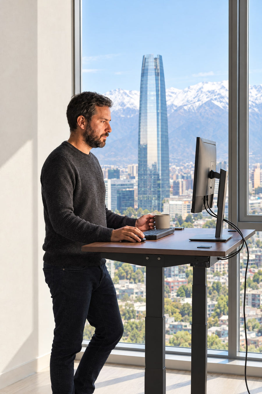 Hombre trabajando de pie en un escritorio eléctrico regulable en altura con vista a la cordillera de Santiago